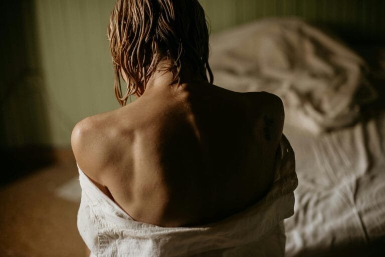 Vrouw met nat haar zit met blote schouders op bed in zacht ochtendlicht, symbool voor kwetsbaarheid en herstel bij fibromyalgie. foto: Natalia Olivera