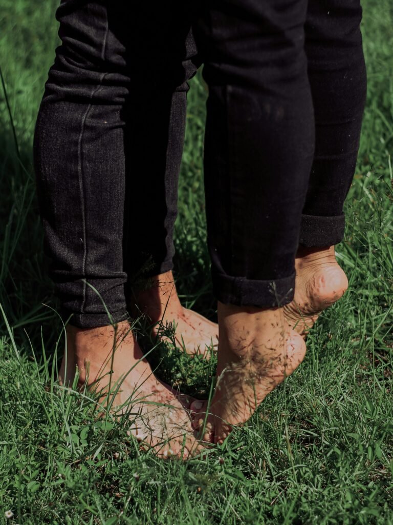Twee mensen staan op blote voeten in het gras, hun voeten dicht bij elkaar, terwijl zonlicht en schaduw over hun enkels en benen vallen, een beeld van aarding, nabijheid en stille aanwezigheid. foto: Carrverg