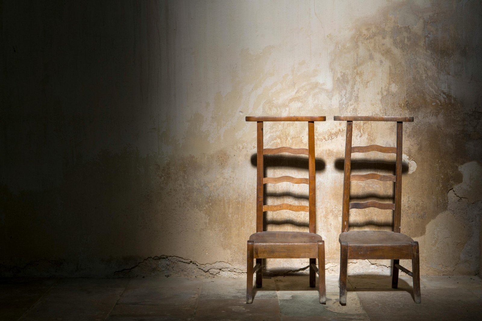 Twee lege houten stoelen naast elkaar tegen een verweerde muur, badend in zacht licht, symbool voor gemis, stilte en verbroken verbinding. foto: Jan Van Der Wolf