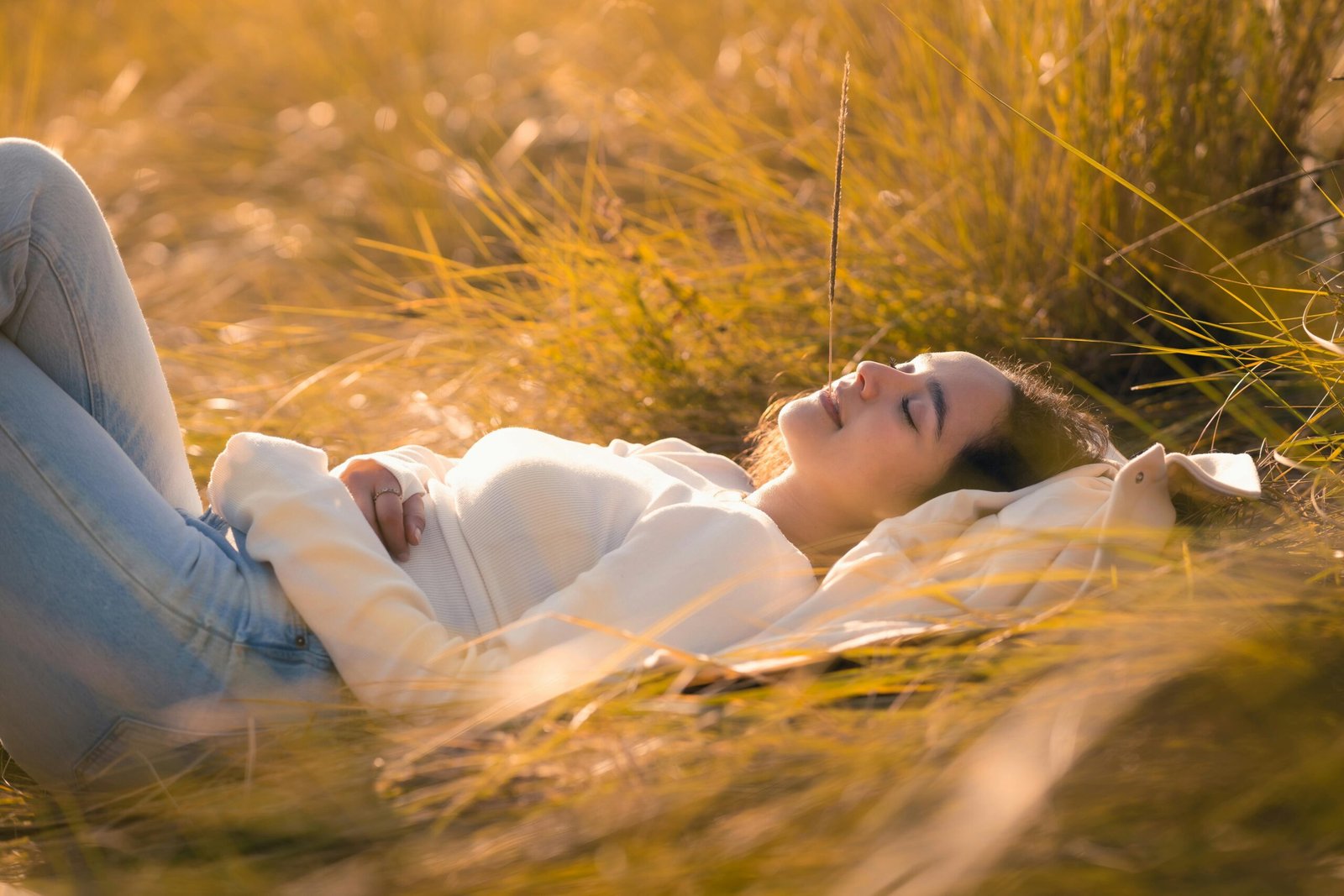 Vrouw ligt ontspannen in warm avondlicht in het gras, ogen gesloten en hand op haar buik, als beeld van zenuwstelselregulatie en herstel na activatie. foto: Julio Lopez