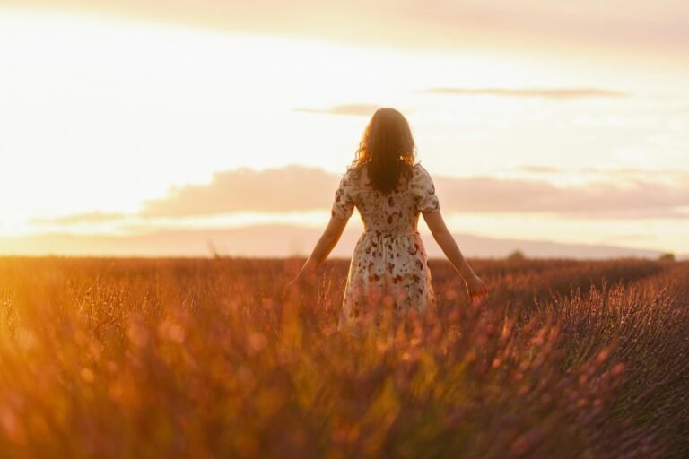 Vrouw staat stil in een veld bij zonsondergang, omringd door hoog gras, symbool voor keuze, timing en het niet forceren van heling bij trauma. foto: Quentin Guiot