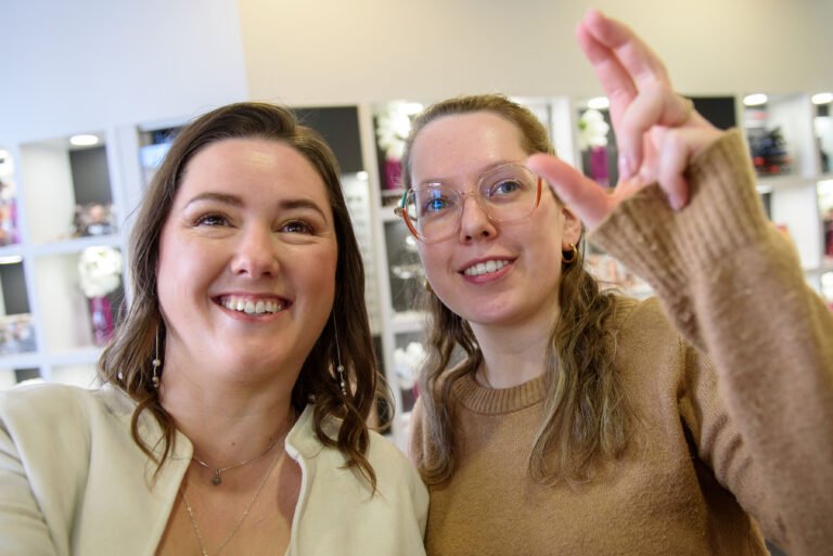 Twee vrouwen glimlachen in een optiekzaak in Wijnegem terwijl één van hen een montuur toont, in een warme en ongedwongen sfeer tussen de brillendisplays. Foto: Studio Tijdloos