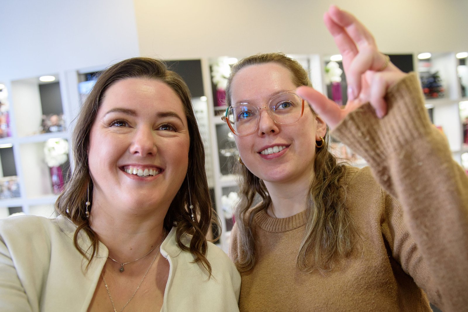 Twee vrouwen glimlachen in een optiekzaak in Wijnegem terwijl één van hen een montuur toont, in een warme en ongedwongen sfeer tussen de brillendisplays. Foto: Studio Tijdloos